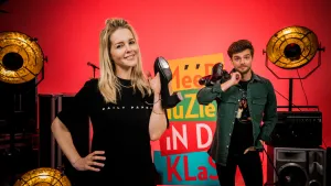 Een foto van Chantal Janzen en Buddy Vedder. Chantal lacht en houdt een zwarte schoen met een hak eronder omhoog, Buddy heeft twee zwarte schoenen vast. Er staan luidsprekers, een radio, en drie draaibare lichten voor op een podium achter ze.
