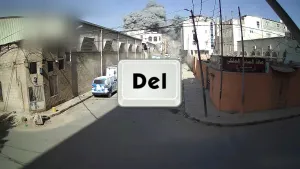 Een foto van een straat in Syrië. Boven de gebouwen achteraan stijgen grijze rookwolken op. Vóór de foto is een getekende toets van een computertoetsenbord te zien. Het is de toets "Del" dat staat voor het Engelse woord Delete, wat "verwijderen" betekent.