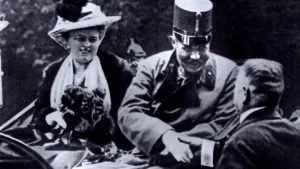 Een zwart-wit foto van de Oostenrijkse kroonprins Frans Ferdinand en zijn vrouw in een rijtuig. Hij draagt een donkere jas en een hoge, ronde hoed met klepje. Hij geeft een man die ernaast staat een hand. Zijn vrouw heeft bloemen vast, het lijken rozen.