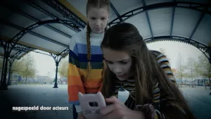 Een foto van twee meisjes buiten onder een hoog afdak. Voor hen staat een mobiele telefoon in een roze hoesje met kattenoren en snorharen. Ze kijken allebei naar het scherm en het rechtmeisje tikt op het scherm. Linksonder staat "nagespeeld door acteurs".