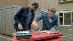 Een foto van Edson en Tim die buiten staan achter een rode tafel. Op tafel staat een grijze bak met zilverkleurig aluminiumfolie erop, ligt een doosje lucifers en een rol aluminiumfolie. Tim houdt een brandend, wit papiertje boven een lichtbruine kaars.