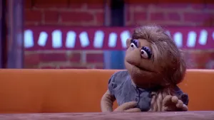 Een foto van pop Vetra op een bruine bank achter een bruine tafel. Vetra heeft een paarse blouse aan, lichtpaarse oogschaduw boven haar ronde ogen en lang, bruin haar. Ze kijkt naar links, naar de niet-zichtbare Opper de Pop.