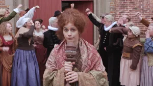 Een foto van een vrouw met een soort microfoon in haar handen. Ze draagt ouderwetse, wijd zittende kleding. Erachter zijn twee donkerrode deuren zichtbaar met aan beide kanten mensen in ouderwetse kleding die naar elkaar roepen en een vuist omhooghouden.