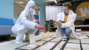 Een foto van iemand in wit veiligheidspak, mondkapje, bril en handschoenen met ervoor rechthoeken van zwart plakband. Er staan bordjes met de cijfers 1, 5 en 30 naast en bij de 1 ligt een boterham. Tim heeft een telefoon vast waarop 25 seconden staat.