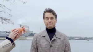 Een foto van Rik van de Westelaken buiten voor een waterweg met boten. Hij kijkt serieus naar de camera in zwarte kleding onder een grijs met bruine jas. Ernaast houden handen een roze varken vast met rood en wit gestipte kleding en een rode haarstrik in.