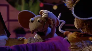 Een foto van pop Ieniemienie met grijze vacht, twee grote, ronde oren en een kronkelende staart. Haar poot ligt op de rand van een kist waar een gestreept en een paars gekleurd kledingstuk aanhangen. Pop Tommie is te zien met zwarte hoed en een nepvogel.