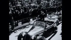 Een zwart-wit foto van de Italiaanse dictator Mussolini die vóór zijn zitting op de achterbank staat in een grote, donkergekleurde auto met opengeklapte bovenkant. Hij draagt een donkergekleurd pak en een pet. Aan de kant staan mensen met vlaggen.