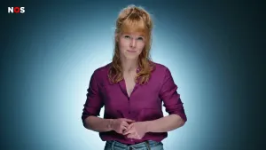 Een foto van een presentator voor een blauwe achtergrond. De presentator draagt een paarse blouse en een stukje van haar blauwe spijkerbroek is ook nog net te zien.