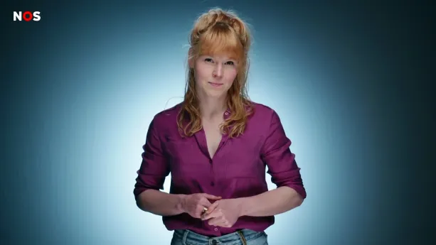 Een foto van een presentator voor een blauwe achtergrond. De presentator draagt een paarse blouse en een stukje van haar blauwe spijkerbroek is ook nog net te zien.