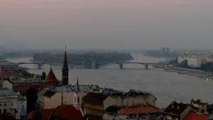 Een foto van de stad Boedapest in Hongarije. Er zijn gedeeltes van gebouwen en huizen te zien aan twee waterkanten en vier verschillende puntdaken van gebouwen. Middenin ligt een waterweg die zich splitst in twee delen en waar een brug overheen loopt.