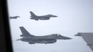 Een foto genomen uit een raam van een vliegtuig, waarin drie gevechtsvliegtuigen of F-16's te zien zijn in de lucht. Twee helemaal en één deels. De F-16's hebben een grijsachtige kleur en een doorzichtige koepel bovenop de voorkant waar de piloot inzit.