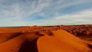 Een foto van een woestijn vol zandheuvels in Egypte. De zandheuvels hebben op de foto een bruine, bijna rode kleur. In de blauwe lucht erboven hangen enkele witte wolken.