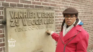 Een foto van Eva die voor een muur staat en naar een rechthoekig monument op de muur wijst. Op het monument staat "Vanhier werden zij weggevoerd - 1 en 2 october 1944". Het is een monument voor alle slachtoffers van de nazi's bij een razzia in Putten.