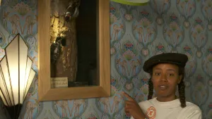 Een foto van presentator Eva. Ze staat in een kamer en wijst naar een foto in een houten lijst. Op die foto zie je twee spechten op een boomstam zitten. 