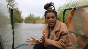 Een foto van Sosha die gehurkt op een boot zit. Rechts van haar hangt een oranje met wit gestreepte reddingsboei en een samengeknoopt touw aan een groene zijkant van stalen buizen. Achter haar zijn het water en de groene struiken ernaast te zien.