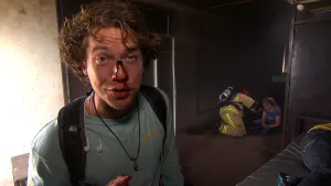 Een foto van Pascal die een nagemaakte wond met rood bloed op zijn neus heeft zitten. Het bloed loopt van bovenaan zijn neus naar zijn neusgaten. Erachter zit een vrouw op de grond die acteert dat ze gewond is naast een brandweerman in brandweerkleding.