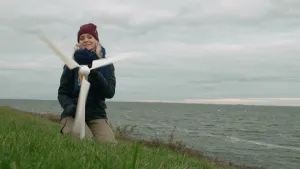 Een foto van presentator Janouk. Ze zit geknield op een dijk achter een schaalmodel van een windmolen. Achter haar zie je water.