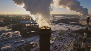Een foto van twee ronde pijpen van een fabriek, die allebei lichtgrijze tot witte rook uitstoten. Op de grond liggen, van veraf zichtbaar, fabrieksgebouwen op een industrieterrein dat bedekt is met een witte laag sneeuw.