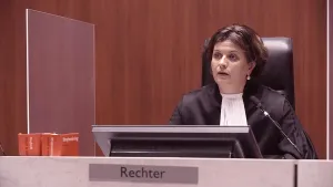 Een foto van een rechter in een zwarte stoel. De rechter draagt een zwart pak met brede mouwen en een witte, rechthoekige doek om haar nek. Vóór de rechter staan een beeldscherm, een microfoon en twee rode wetboeken en vooraan hangt een bordje "Rechter".