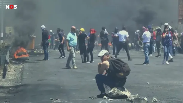 Een foto van een weg met mensen, zwarte rookwolken erboven en autobanden die in brand staan. De meeste mensen hebben een mondkapje of doek voor hun hoofd en een paar hebben een autoband vast. Vooraan zit één man op een steen en kijkt naar achteren. 