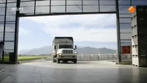 Een foto van een vrachtwagen die een grote loods inrijdt. Je ziet het beeld vanuit de loods gezien. 