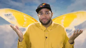 Een foto van Edson die in de camera kijkt en zijn handen met de handpalmen omhoog houdt, om te laten zien dat hij het niet weet. Hij heeft een gele blouse aan en in de achtergrond zijn, precies boven zijn schouders, gele vleugels van een vlinder te zien.