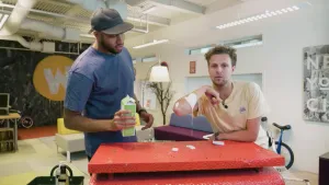 Een foto van Edson en Tim achter een rode tafel. Edson heeft een pak yoghurt in zijn hand. Op de onderarm van Tim ligt een klodder yoghurt, dat van zijn arm afloopt op de tafel. Ze testen of de yoghurt helpt tegen jeuk van brandnetels op hun huid.