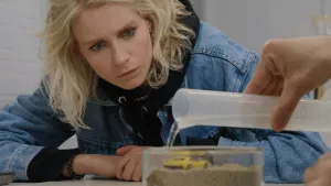 Een foto van presentator Janouk. Ze kijkt aandachtig naar een doorzichtige bak. Daarin zit zand met daarop een modelautootje. Iemand giet water in die bak. 