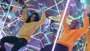 Een foto van presentator Sosha. Ze is samen met een man aan het dansen in een ruimte met veel spiegels op de achtergrond. Je ziet ook veel kleuren in die spiegels weerkaatst.