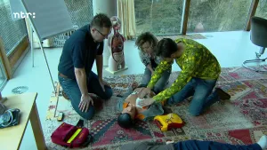 Een foto van drie mensen die geknield bij een plastic pop zitten. Eén van de mensen drukt met de handen op het linkerdeel van de borstkas van de pop. Ze doen na hoe iemand gereanimeerd moet worden, als het hart van iemand stopt met kloppen. 