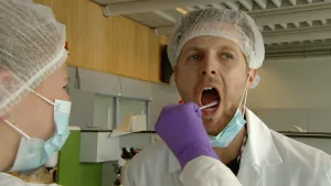 Een foto van Tim in een witte jas, met een wit haarnetje op zijn hoofd en een lichtblauw mondkapje, dat onder zijn nek hangt. Wies draagt dezelfde kleding en steekt een in plastic gehandschoende hand in de wijdopen mond van Tim, om speeksel af te nemen.