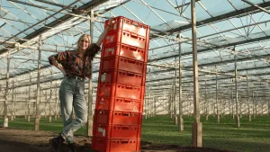 Een foto van presentator Janouk in een grote kas. Ze staat naast een stapel rode kratten die hoger is dan zijzelf. Ze leunt op het bovenste krat.