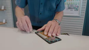 Een foto van een man waarvan het gezicht niet te zien is, die achter een tafel staat, een mobiele telefoon uit elkaar haalt en een pincet vastheeft. Te zien zijn het groene moederbord waarop alle onderdelen zitten en de grijze batterij van de telefoon.