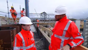 Een foto van Edson en Pepijn in een oranje vest met zilverkleurige strepen erop, met doorzichtige veiligheidsbril en met een witte helm. Ze staan op een bouwplaats op een ondergrond van stalen rekken. Achter hen staan drie mannen op een hoger gelegen rek.