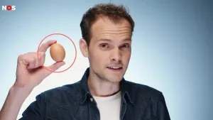 Een foto van een presentator die tussen duim en wijsvinger van zijn rechterhand een bruin ei heeft van een kip. Vóór de foto is om het ei een rode cirkel getekend. De achtergrond van de foto is blauw.