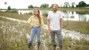 Een foto van twee mensen met laarzen aan die naar de camera kijken. Ze staan op een natte grond met groen gras, omdat de rivier rechts ernaast buiten haar oevers is getreden, is overstroomd. Achteraan, aan de andere kant van de rivier, staan bomen.