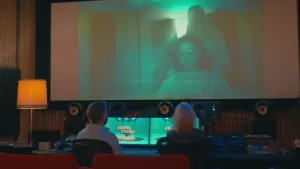 Een foto van prof en Janouk achter een tafel met daarop twee beeldschermen, apparaten met knopjes en schuifjes en luidsprekers. Op de muur erboven hangt een scherm met daarop een jongen die zit en een meisje dat erachter staat in een donkere kamer.