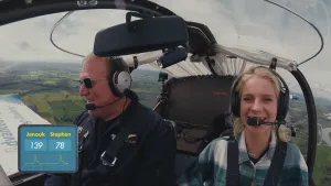 Een foto van een tweepersoons vliegtuig in de lucht boven een groen landschap met daarin piloot Stephen en Janouk. Linksonder is een hartslagmeter te zien, bij Janouk staat de waarde 139, bij Stephen 78. Ze lachen en dragen een koptelefoon met microfoon.