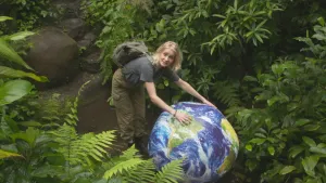 Een foto van presentator Janouk. Ze staat in een nagemaakt stukje tropisch regenwoud en rolt een grote bal voor zich uit. Die bal ziet er uit als de Aarde.