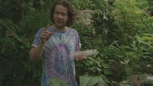 Een foto van presentator Pascal. Hij staat in een vlindertuin en legt iets uit aan de camera. Hij heeft in een hand een plastic bakje vast.