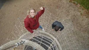 Een foto van presentator Janouk. Ze staat aan de buitenkant van een klimtoren. Het beeld is van boven naar beneden gefilmd. Onder haar zie je een vangnet en een man die daar op staat.