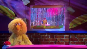 Een foto van pop Opper de Pop die op de foto staat bij een rode drukknop. Erachter staat de pop Kip Tandoori, een kip met een rode kam op het hoofd, achter een rechthoekige opening in de muur. Vóór de kip staat een zoutvaatje van doorzichtig glas.