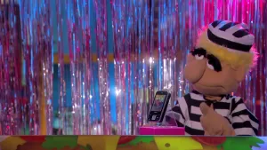 Een foto van de pop Joris Boef die dikke, zwarte wenkbrauwen heeft. De pop draagt een pak en een pet met witte en zwarte strepen, zoals boeven vroeger droegen in de gevangenis. Hij wijst met zijn vinger naar een zwarte mobiele telefoon op de boterhamband.