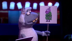 Een foto van pop Stella Schaap. De pop heeft een bord vast met een getekende oude vrouw met grijs haar erop die een groene jurk aanheeft. De pop heeft een witte vacht, de kop en poten zijn grijs, de ogen rond en de vooruitstekende tanden geel van kleur.