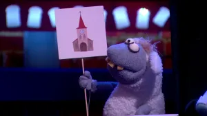 Een foto van pop Stella Schaap. De pop heeft een bord vast met een getekende kerk erop, een groot gebouw met een rood dak waar een klok onder hangt. De pop heeft een witte vacht, de kop en poten zijn grijs, de ogen rond en vooruitstekende, gele tanden.