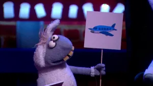 Een foto van pop Stella Schaap. De pop heeft een bord met een getekend blauw vliegtuig erop vast en kijkt ernaar. De pop heeft een witte vacht op haar rug en oren, een grijze vacht bij kop en poten, ronde ogen en vooruitstekende, gele tanden.