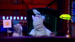 Een foto van pop Stella Schaap die een bord omhoog houdt met een getekende, oranjegele zon erop. Naast haar zit pop Nico de Neushoorn op een groene stoel met zijn bril op en op de tafel ervoor ligt een opengeklapt boek en staat een theekopje.