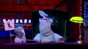 Een foto van pop Stella Schaap die een bord omhoog houdt met een getekende, rode papegaai erop met gele en blauwe vleugels. Naast haar zit pop Nico de Neushoorn op een stoel en op de tafel ervoor zijn een opengeklapt boek en een theekopje te zien.