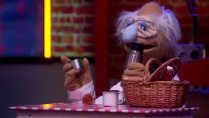 Een foto van pop professor Bolleboos met grijs haar, rimpelig gezicht en lichtblauwe neus. Hij heeft een thermosfles vast om drinken warm of koud mee te houden. Voor hem op tafel staat een picknickmand met flesje, een klein potje jam en een witte beker.