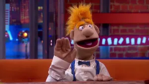 Een foto van pop professor Adieu die op een bruine bank aan een bruine tafel zit. De pop zwaait naar het niet-zichtbare publiek. Hij heeft oranje haar, een witte blouse met blauwe strik aan en blauw met wit gestreepte bretels om zijn schouders.